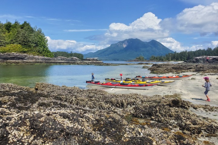 Colorful kayaks on a beach with mountains and forests in the background, under a partly cloudy sky.