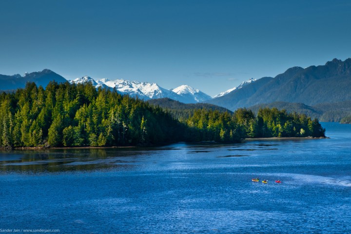 Kayakers in blue water with forested islands and snowy mountains under a clear blue sky.