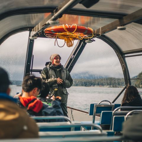 Guide speaking to tourists inside a boat with large windows, life ring above, and a scenic lake view.