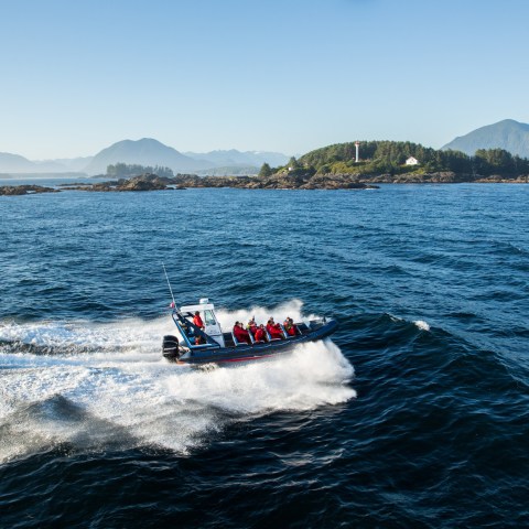 Speedboat with people in red gear on ocean near island with lighthouse, mountains in background.