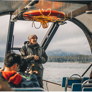 Person speaking to a group inside a boat with mountains and water in the background.