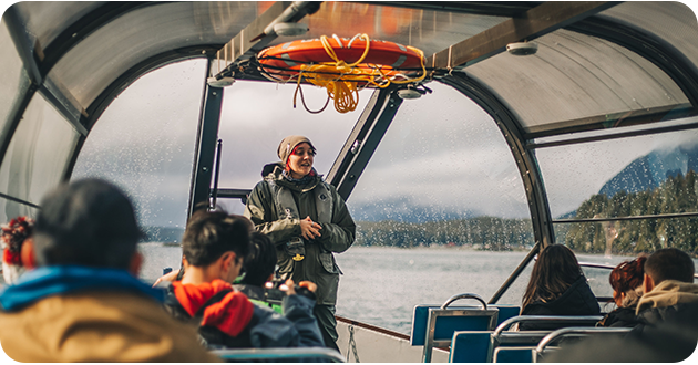 Person speaking to a group inside a boat with mountains and water in the background.