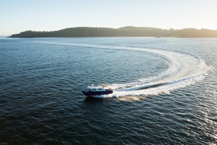 Boat making a large circular wake in calm ocean near forested island at sunset.